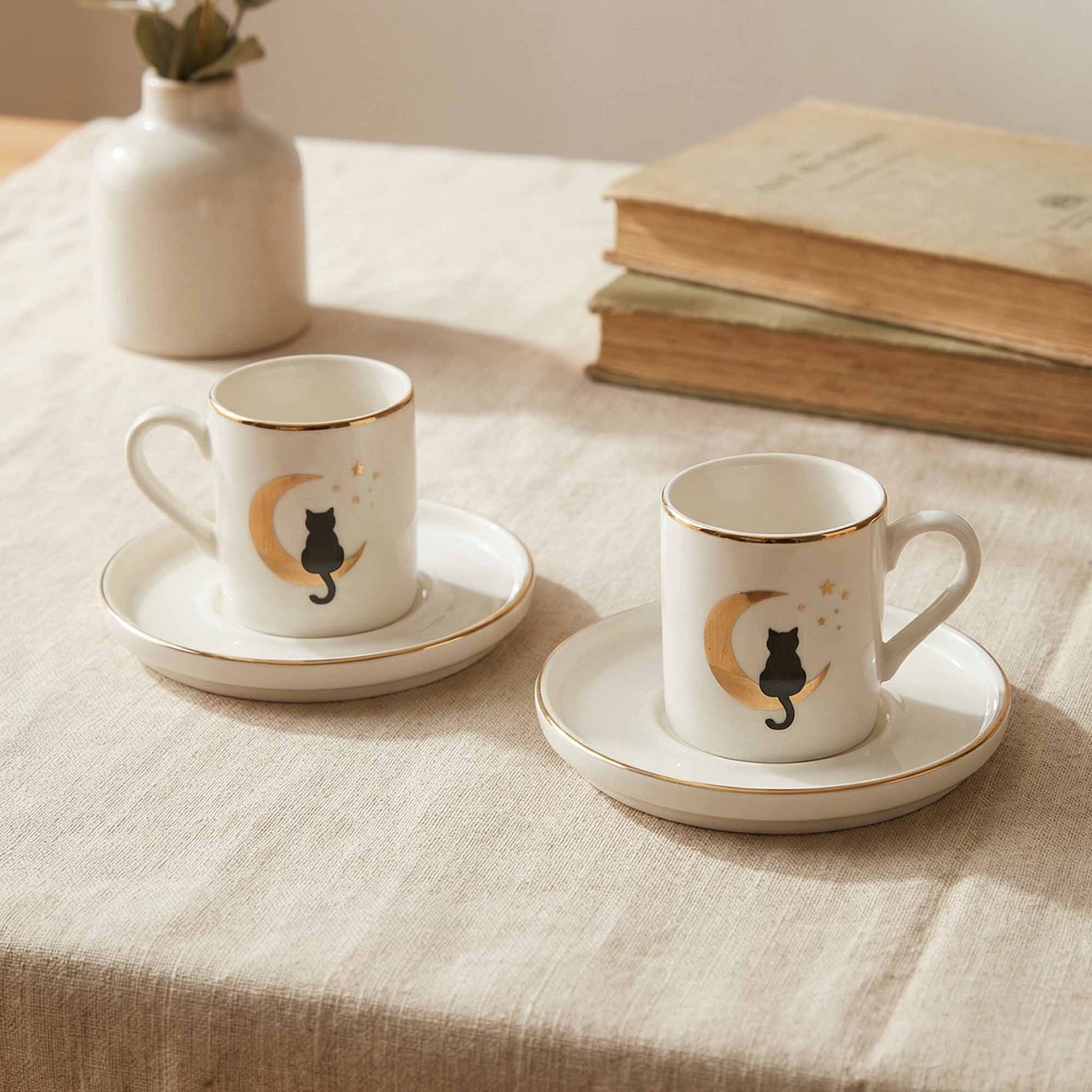 Two white coffee cups with black cat designs on a beige tablecloth with books and a vase in the background.