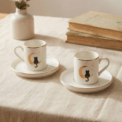 Two white coffee cups with black cat designs on a beige tablecloth with books and a vase in the background.
