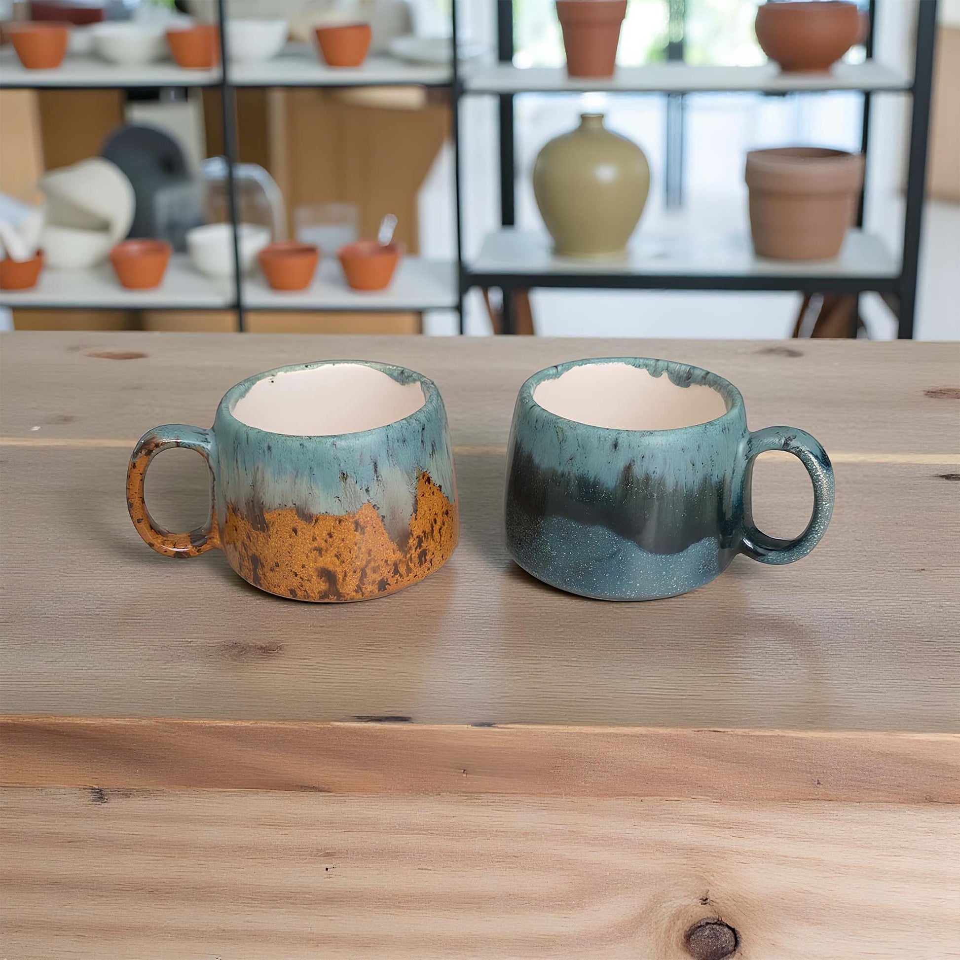 Two ceramic mugs with a rustic finish on a wooden surface, with shelves of pottery in the background.