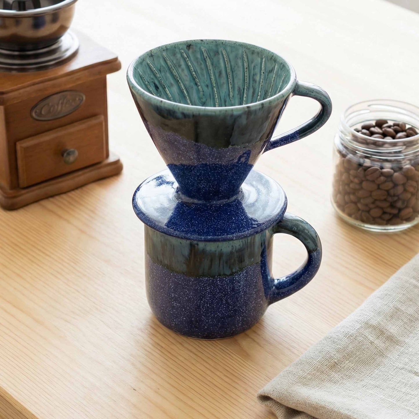 Ceramic coffee dripper with blue and green design on a wooden surface with coffee beans and a grinder.