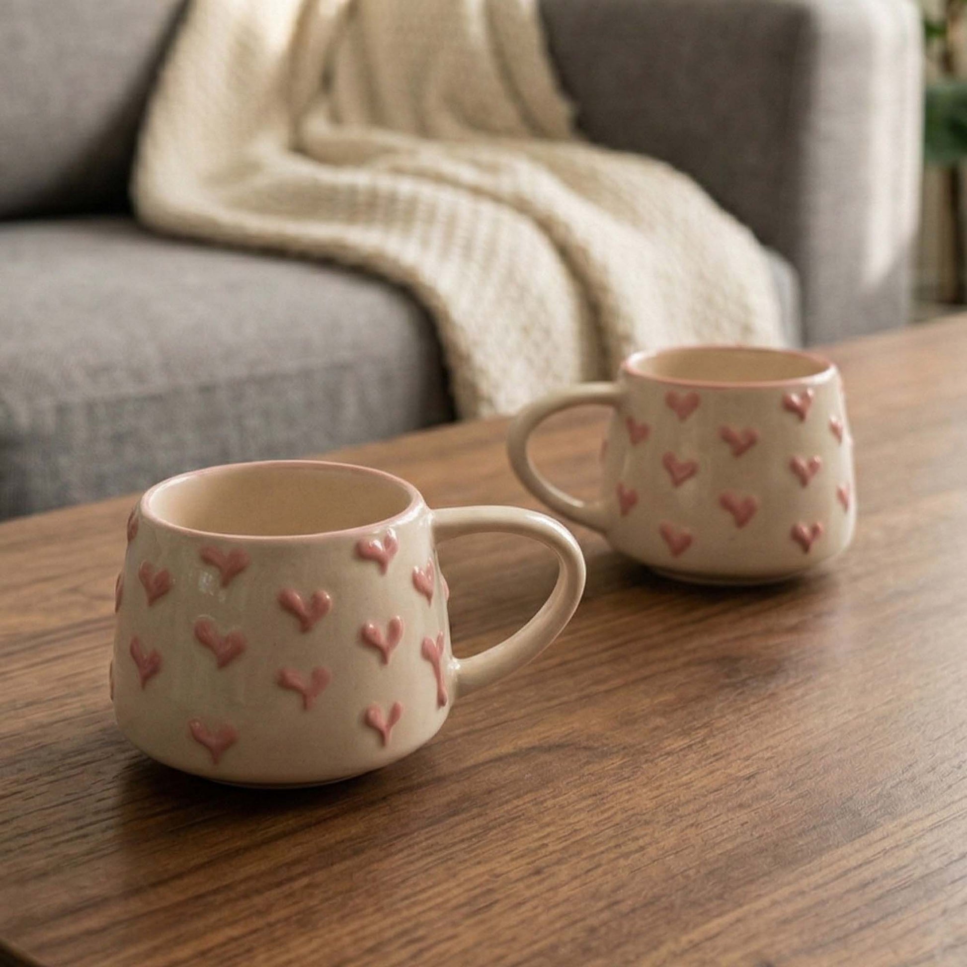 Two ceramic mugs with heart patterns on a wooden table, with a blurred background of a sofa and blanket.