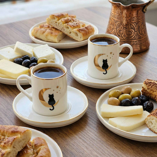 Two cups of coffee with saucers on a wooden table with plates of bread, cheese, and olives.