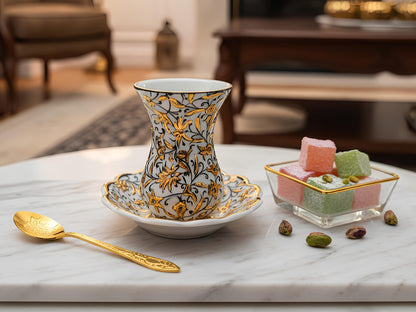 Decorative teacup with gold patterns on a marble surface, accompanied by a spoon and small desserts.