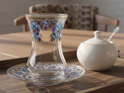 Decorative glass with floral patterns on a wooden table with a teapot in the background.
