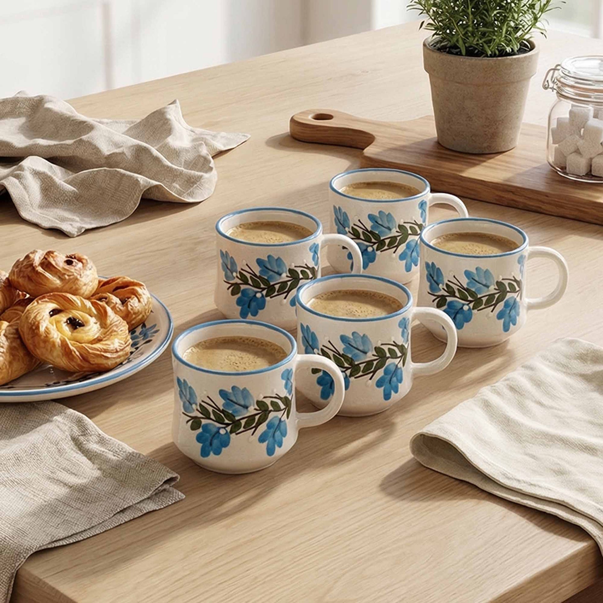 Set of white mugs with floral designs filled with a beverage on a wooden table with pastries and a plant.