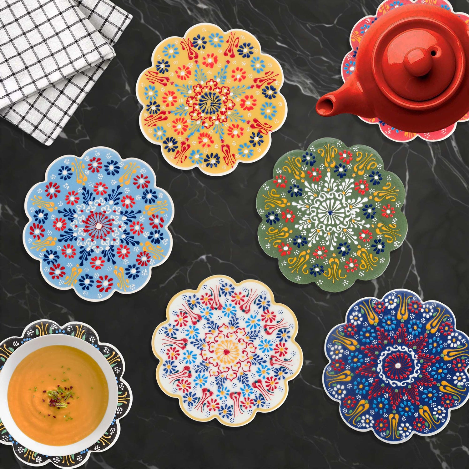 Colorful floral trivets on a dark surface with a red teapot and checkered cloth.