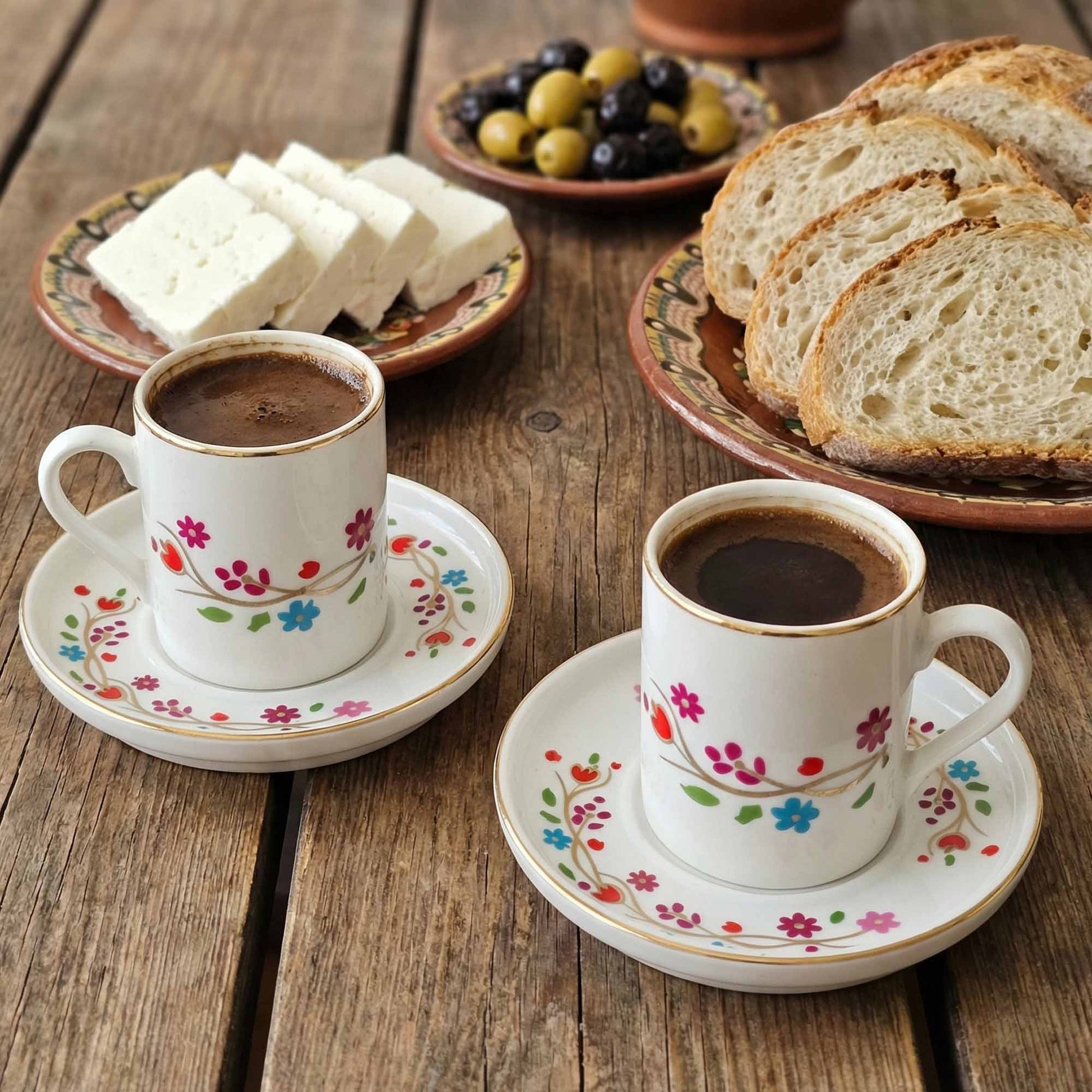 Two cups of coffee with floral designs on saucers, accompanied by bread and olives on a wooden table.