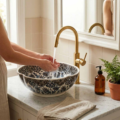 Person washing a floral-patterned bowl under a gold faucet in a bathroom.